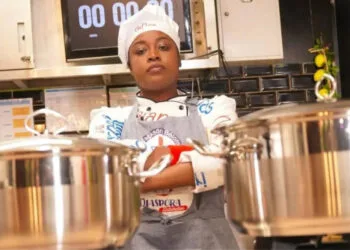 La cheffe haïtienne Leen Excellent prépare un plat traditionnel haïtien lors de son marathon culinaire de 216 heures, entourée d’ustensiles et d’ingrédients locaux.