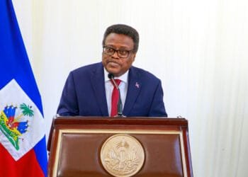 Fritz Alphonse Jean au micro, devant un drapeau haïtien en berne, illustrant l’incertitude et la lourde responsabilité face à la crise nationale.